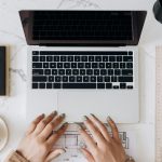 Top view of a stylish home office desk with a laptop, planner, and coffee cup, showing hands on a blueprint.